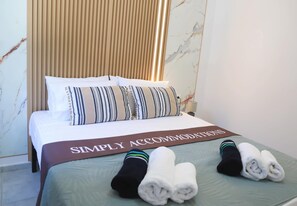 Ein ordentlich bezogenes Bett mit einem 'SIMPLY ACCOMMODATIONS' Schild, gestreiften Kissen und aufgerollten Handtüchern auf einer grünen Tagesdecke.