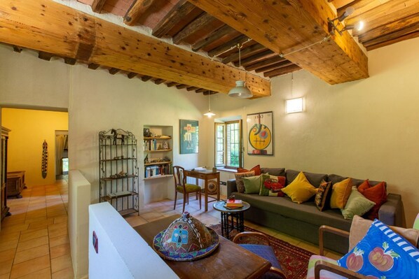 A cozy living room with a wooden ceiling, a sofa with colorful cushions, a bookshelf, and a dining area with chairs.