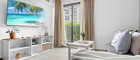 Ett modernt hotellrum med en stor platt-TV, en balkong med utsikt över havet, en vit soffa och ett litet bord med en vas.