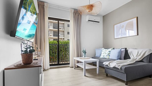 A modern living room with a grey sofa, a white coffee table, a flat-screen TV, and a balcony with a view of buildings and greenery. - NOK Cozy 1BR Retreat With Pool Near Punta Cana Beaches (Punta Cana)