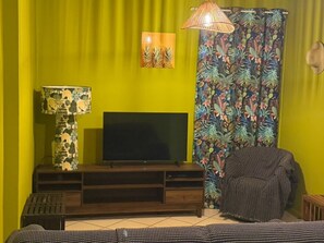 Una habitación con televisión sobre un soporte de madera, una lámpara estampada, una silla y una cortina con motivos tropicales.