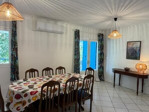 Un comedor con una mesa cubierta con un mantel estampado de frutas, rodeada de sillas de madera. Hay dos lámparas colgantes, un ventilador de techo y un cuadro en la pared.
