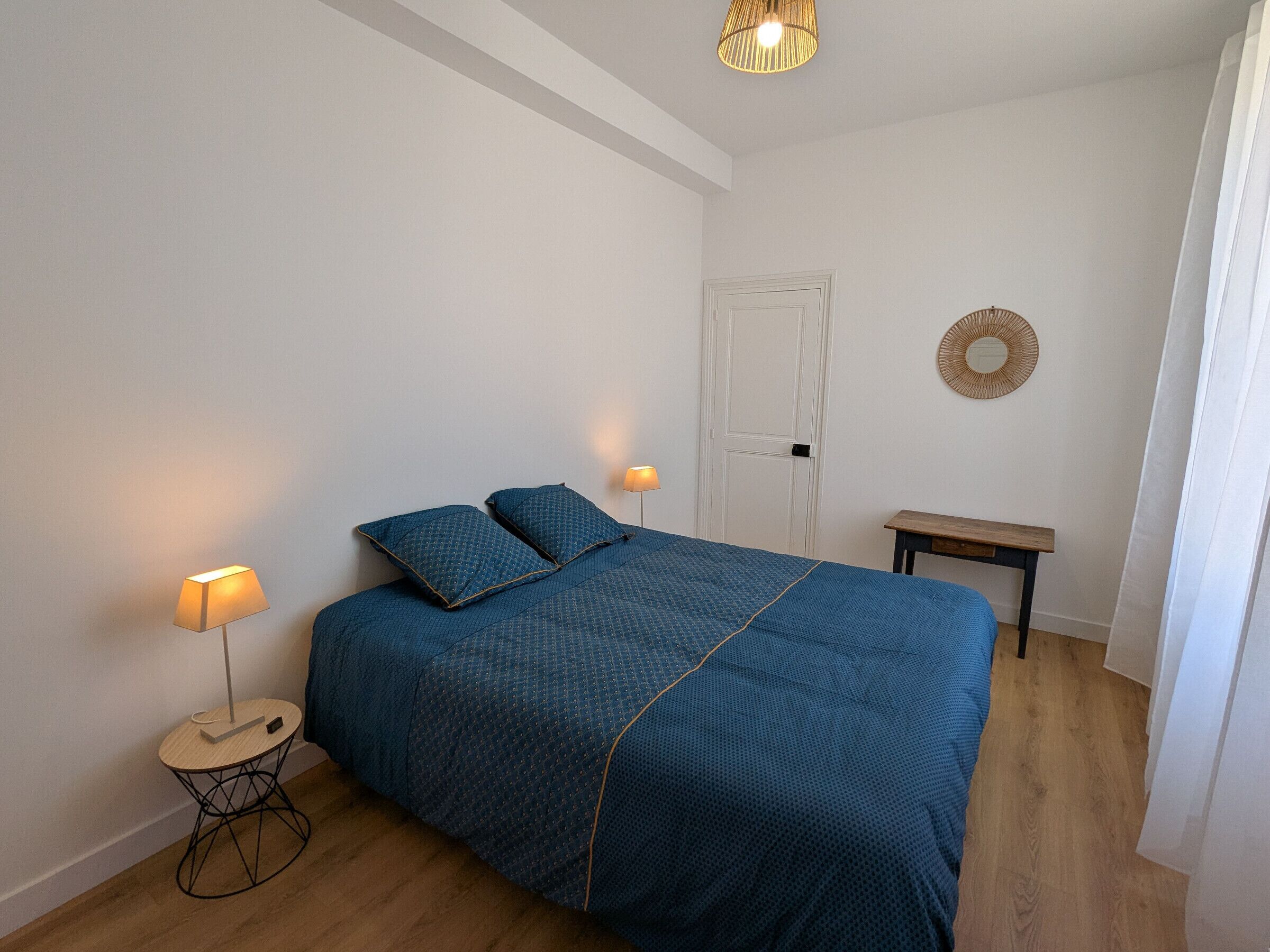 Un dormitorio con una cama, dos lámparas de noche, un espejo redondo, una mesita de madera y una puerta.