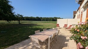 Eine Terrasse im Freien mit Tisch und Stühlen, ein Rosengarten und Blick auf ein Feld mit Bäumen.