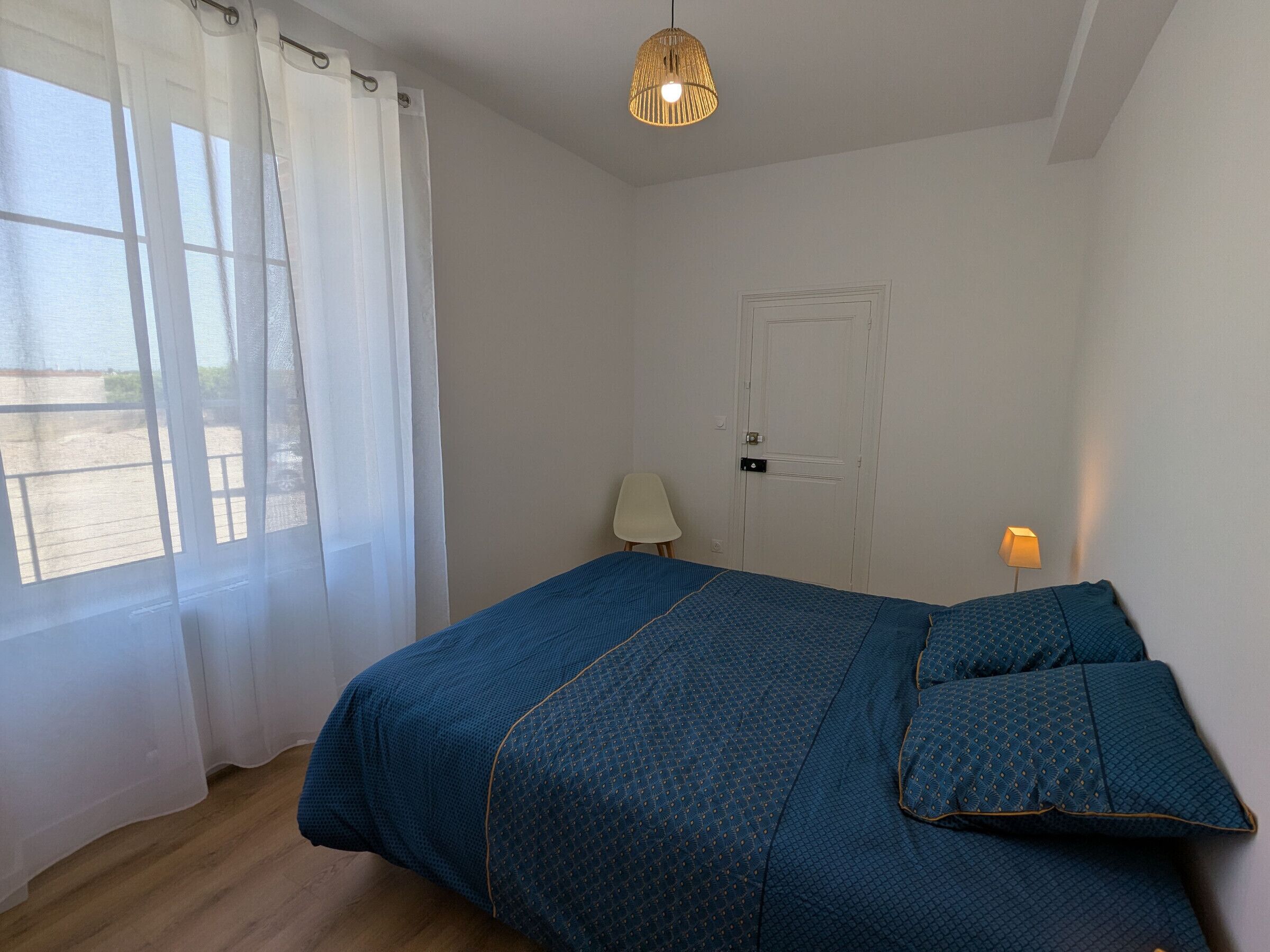Un dormitorio con cama, puerta, silla, lámpara y ventana con cortinas.