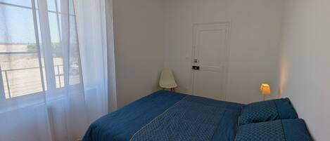 Un dormitorio con cama, puerta, silla, lámpara y ventana con cortinas.