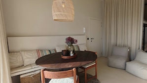 Um quarto aconchegante com uma mesa redonda de madeira, um sofá com almofadas estampadas, uma luminária pendente de vime e vista para uma varanda.
