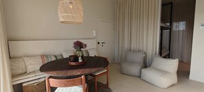 Ein gemütliches Zimmer mit einem runden Holztisch, einer Couch mit gemusterten Kissen, einer Weidenpendelleuchte und einem Blick auf einen Balkon.
