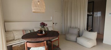 A cozy room with a round wooden table, a sofa with patterned cushions, a wicker pendant light, and a view of a balcony.