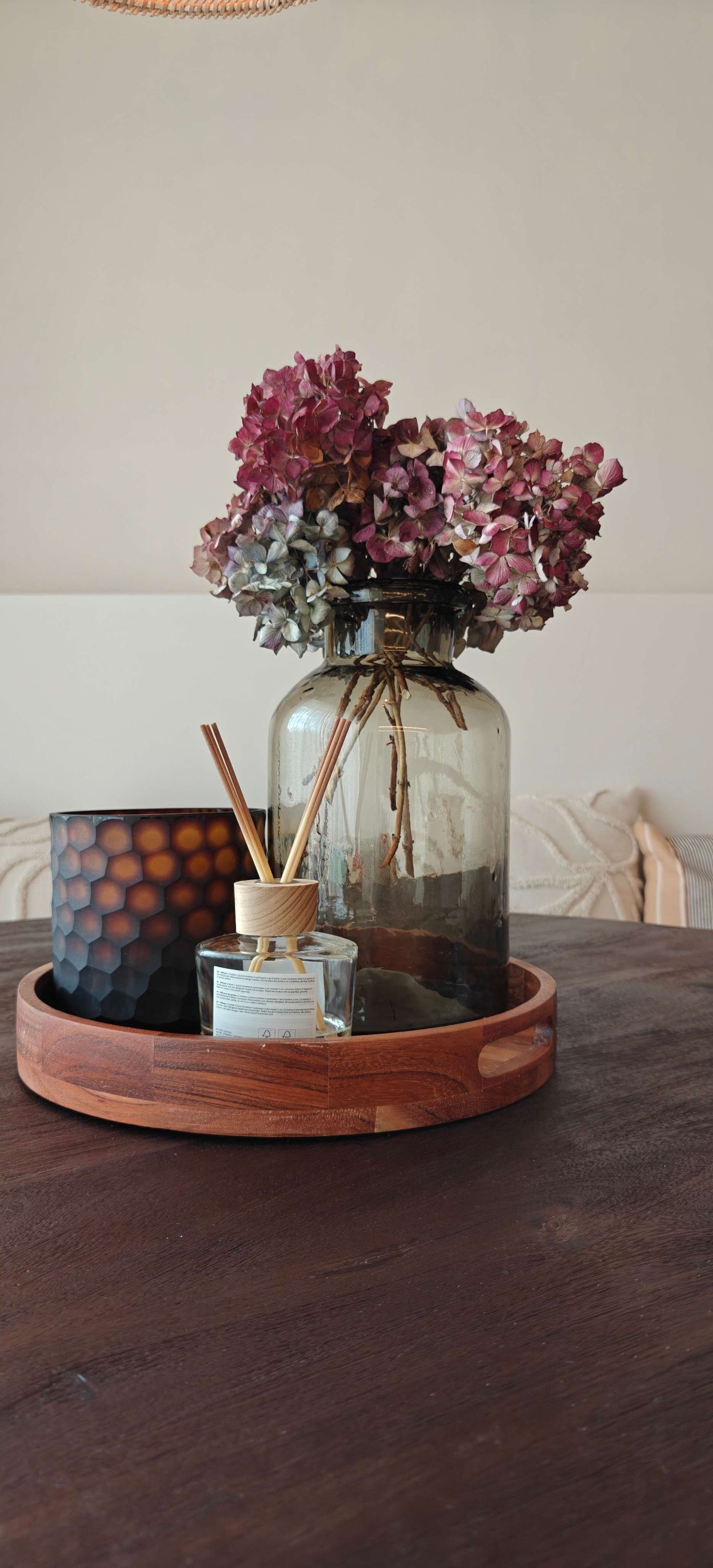 A wooden tray with a glass vase containing dried flowers, a hexagonal candle, and a small bottle with a label.