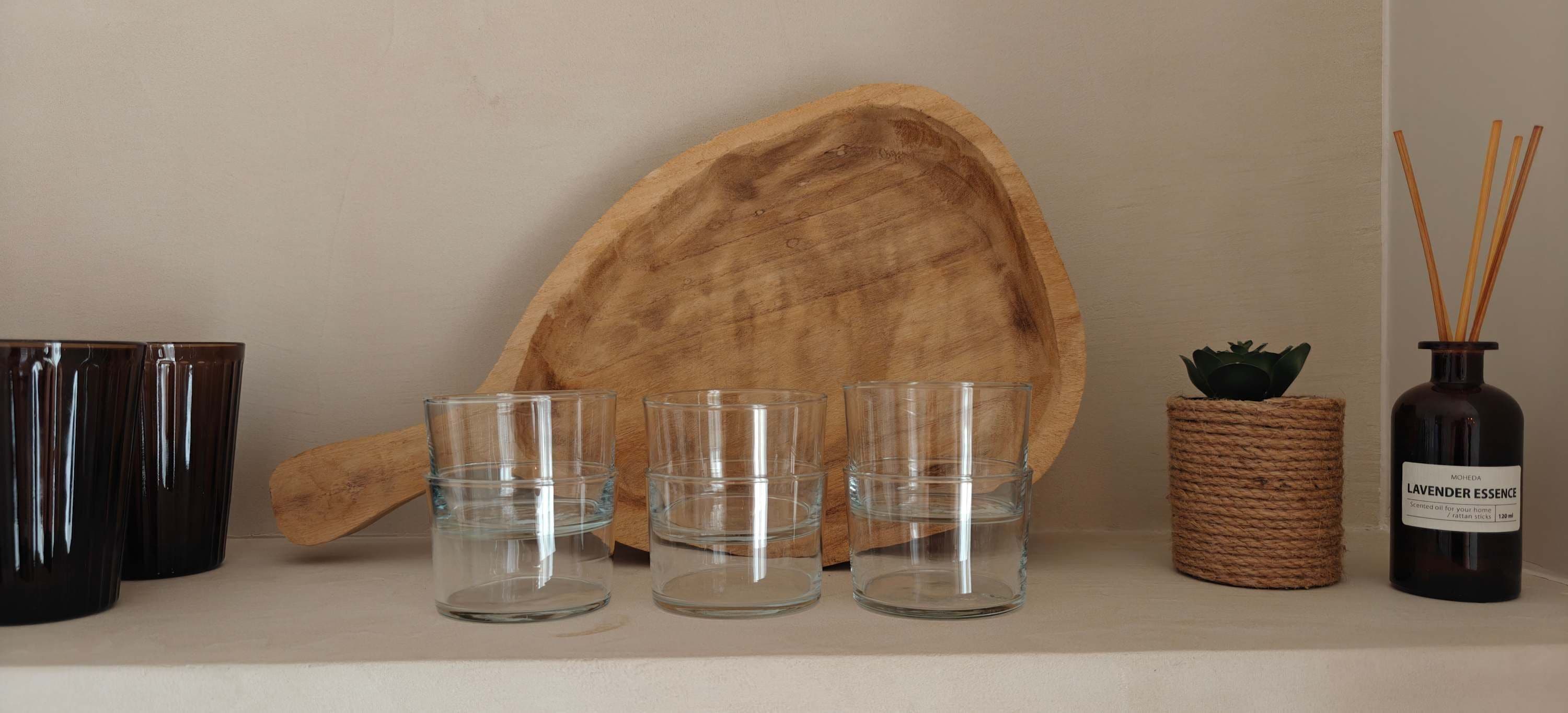 A shelf with four clear glasses, a wooden cutting board, a woven basket with a plant, and a bottle of lavender essence.