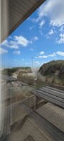 A view from a window overlooking a beach with a wooden bench, a metal fence, and a sandy area with grass.