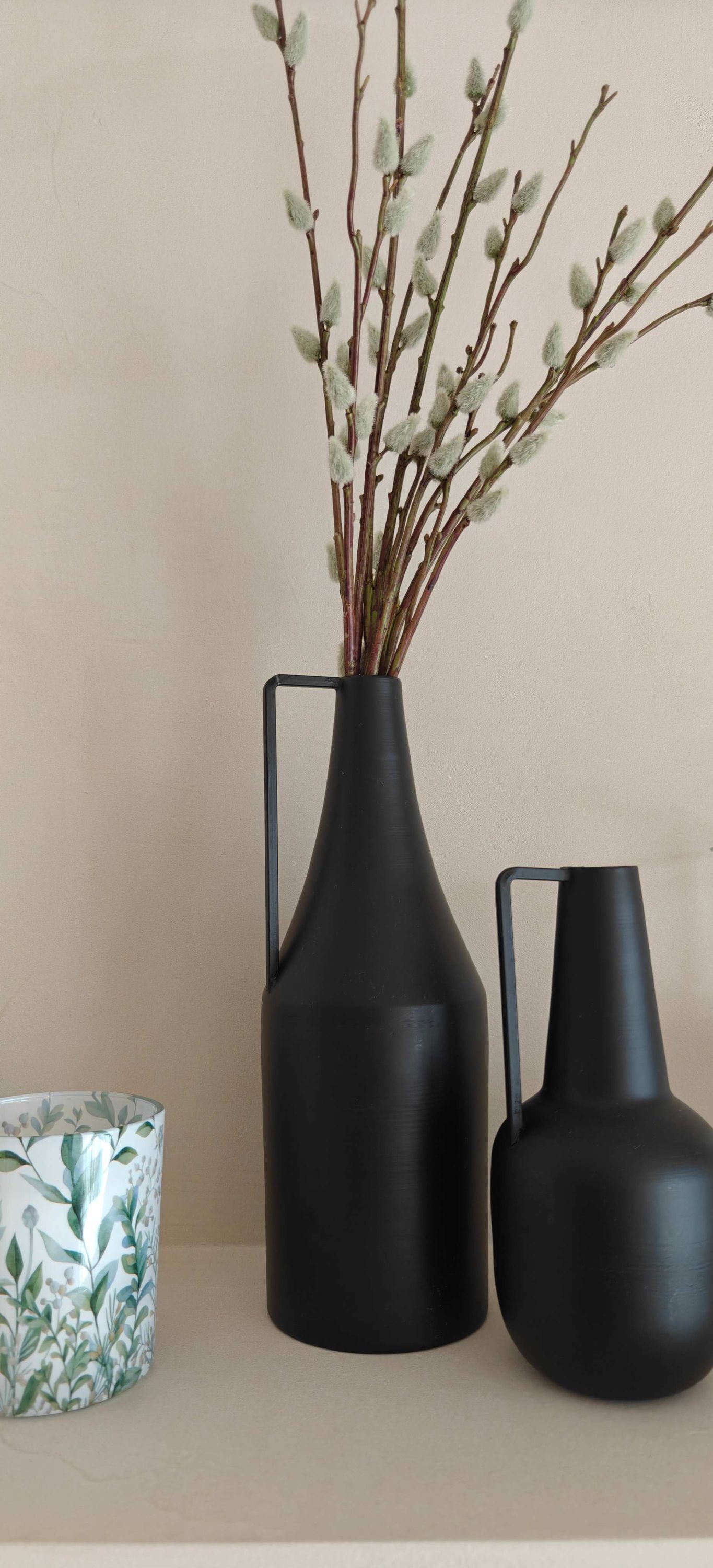 A black vase with branches and leaves, a smaller black vase, and a patterned cup on a shelf.