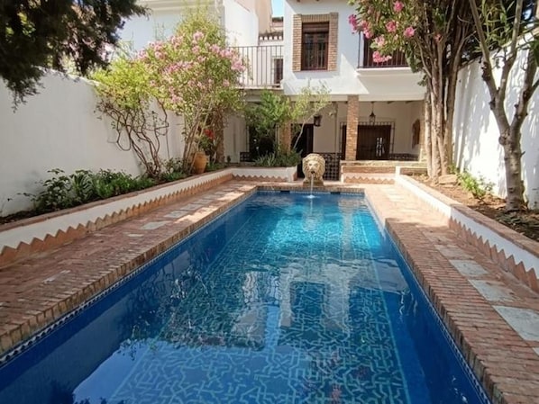 Une piscine rectangulaire avec une fontaine, entourée d’un bâtiment blanc doté de balcons et de plantes en pot.