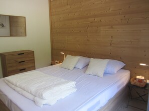 Ein ordentlich bezogenes Bett mit Kissen, ein Holz-Kopfteil, ein Nachttisch mit Lampe und eine Holzkommode.