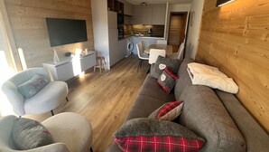 A modern living room with a sofa, chairs, a TV, a kitchen area, and a wooden wall. - Comfortable duplex - 6 guests (Châtel)