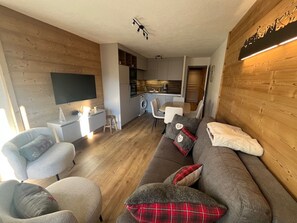 A modern living room with a sofa, chairs, a TV, a kitchen area, and a wooden wall.