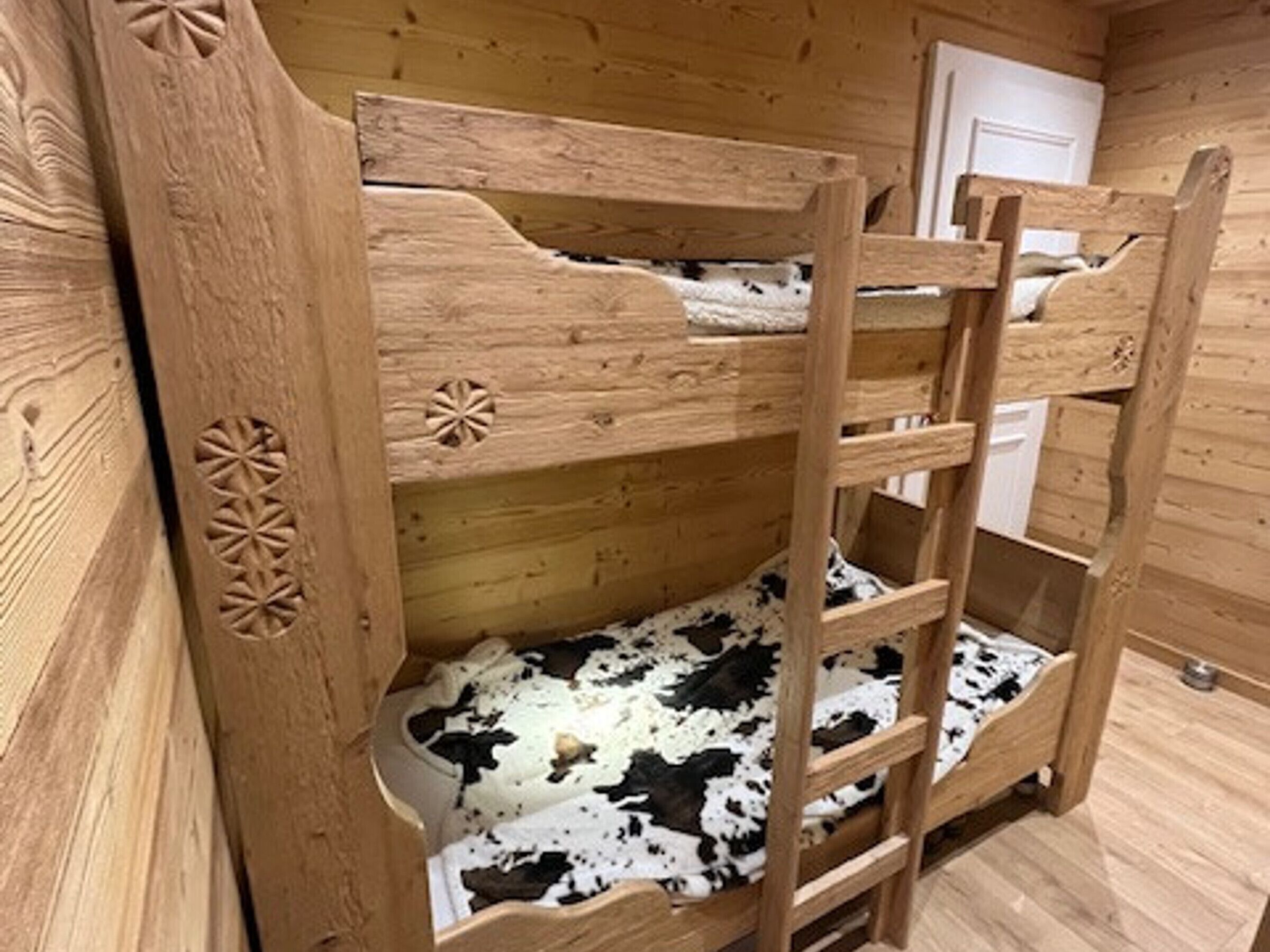Un lit superposé en bois, avec un matelas à motifs de vaches, une échelle et une porte, dans une pièce aux murs en bois.