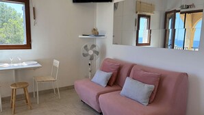 Een kamer met een roze bank, een kleine tafel met glazen, een ventilator en een aan de muur bevestigde televisie.