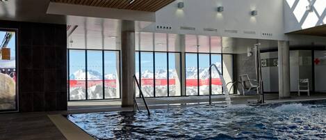 An indoor swimming pool with a view of snow-capped mountains, large windows, and a wooden ceiling.