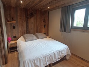 Ein Schlafzimmer mit Holzboden, einem Bett mit weißer Bettwäsche, einem Nachttisch mit Lampe und einem Fenster mit Vorhängen.