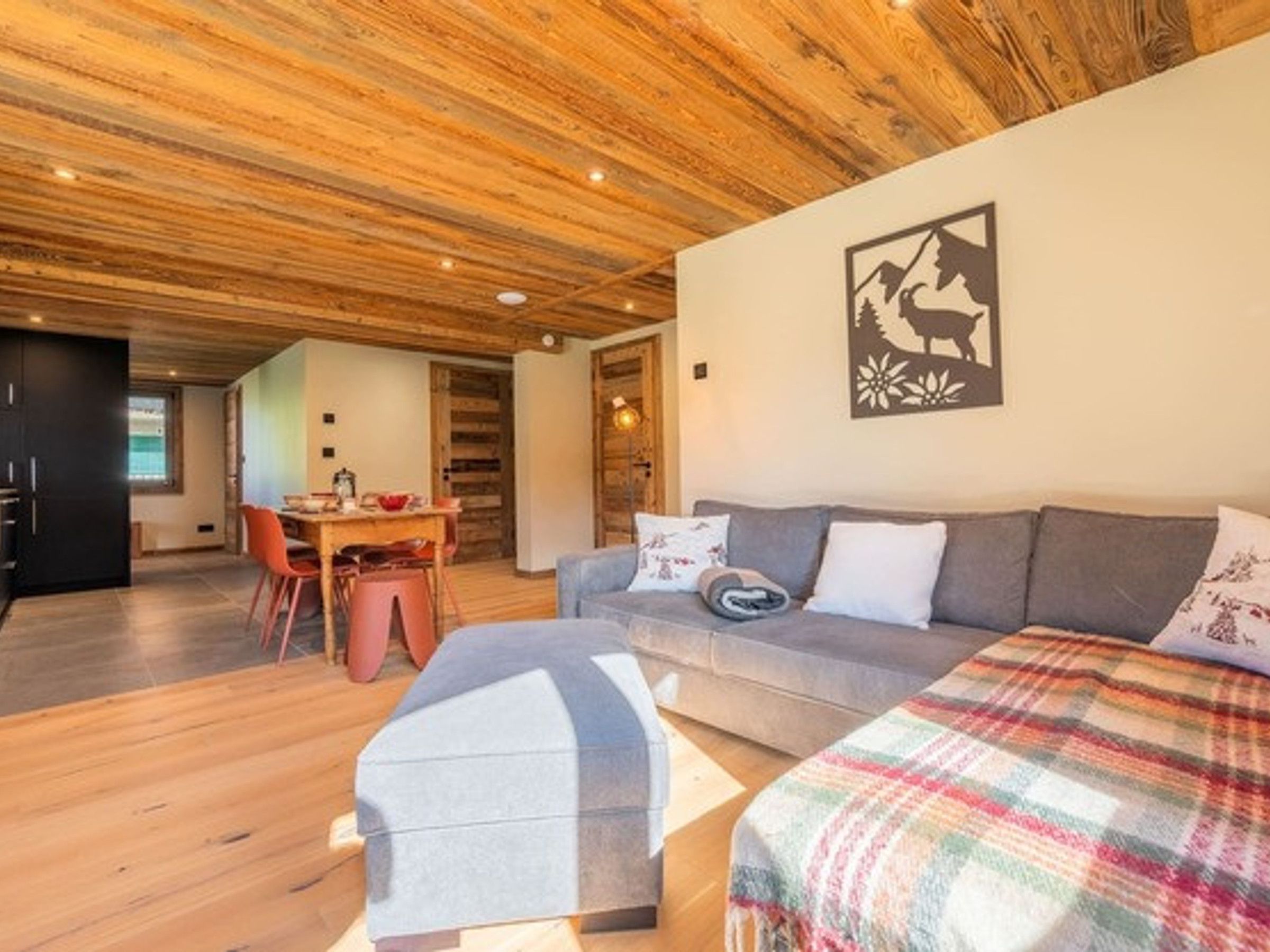 A cozy living room with a wooden ceiling, a sofa, a dining table with chairs, and a wall-mounted artwork.