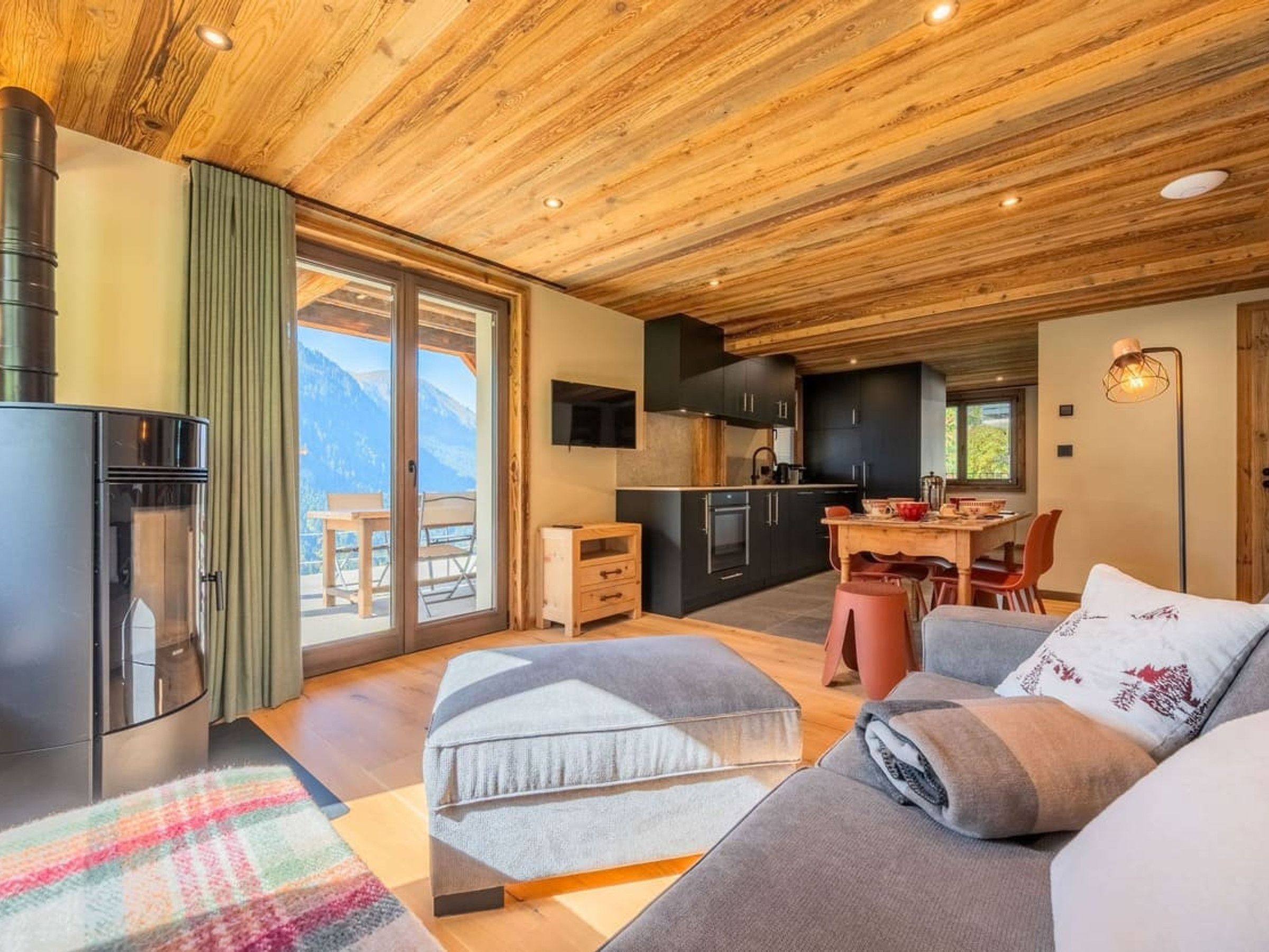 A modern interior with a wooden ceiling, a kitchen area, a dining space, and a sofa with a view of mountains.