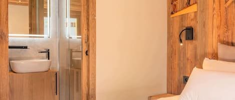 A bedroom with a wooden wall, a bed, a nightstand, a bedside table, a mirror, and a bathroom with a sink and a shower.