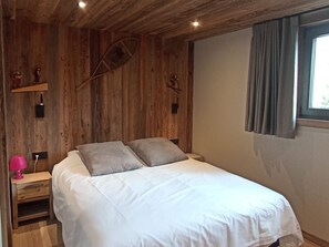Ein Zimmer aus Holz mit einem Bett, einem Nachttisch, einer Lampe und einem Fenster mit Vorhängen.