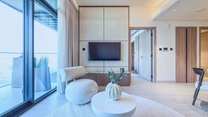 Ein modernes Wohnzimmer mit großem Fernseher, bequemen Sitzmöglichkeiten, einem runden Couchtisch und Blick durch große Fenster.