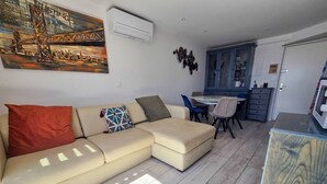A living room with a beige sofa, a dining table with chairs, a kitchen area, and a painting on the wall. - Apartment with AC, port view - 4 guests (Cap d'Agde)