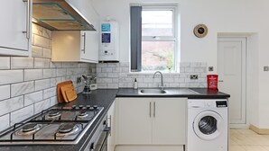 A compact kitchen with a gas stove, a built-in washing machine, a window with a view, and a clock on the wall. - Perfect Booking 4 Bed Highfield House w 4 Baths (Sheffield)