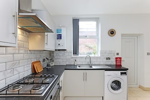 A compact kitchen with a gas stove, a built-in washing machine, a window with a view, and a clock on the wall. - Perfect Booking 4 Bed Highfield House w 4 Baths (Sheffield)