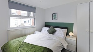 A bedroom with a bed, a nightstand, a lamp, a window with blinds, a radiator, and a framed picture on the wall. - Perfect Booking 4 Bed Highfield House w 4 Baths (Sheffield)