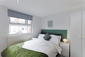 A bedroom with a bed, a nightstand, a lamp, a window with blinds, a radiator, and a framed picture on the wall. - Perfect Booking 4 Bed Highfield House w 4 Baths (Sheffield)