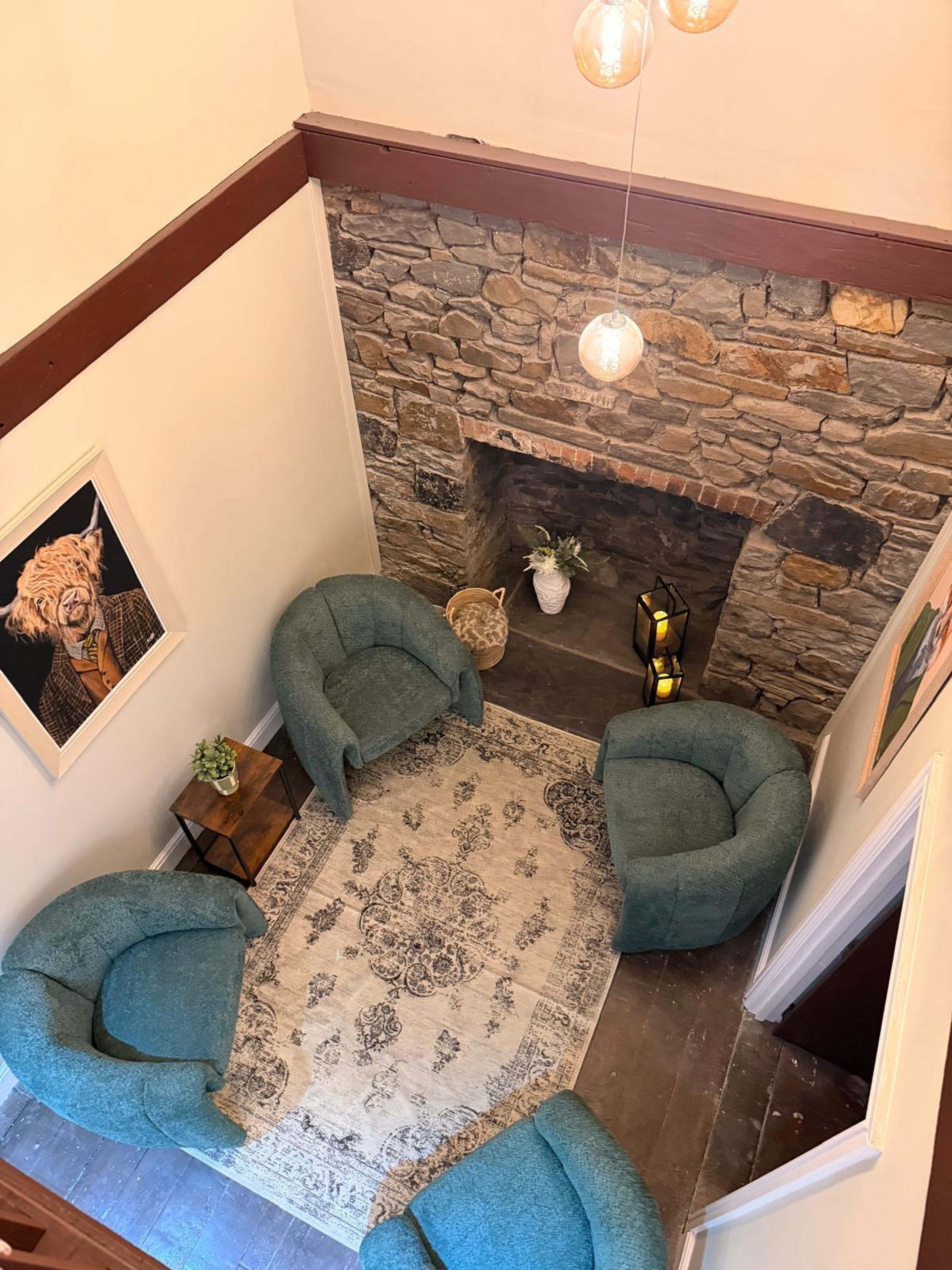 Una sala de estar acogedora con una chimenea de piedra, dos sillones verdes, un tapete estampado y un cuadro en la pared.