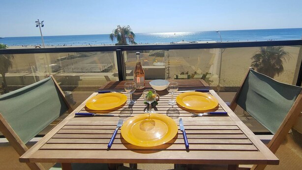 A table set for two with yellow plates, a bottle, and cutlery on a balcony overlooking a beach. - Studio sea view, parking and air conditioning (Canet-Plage)