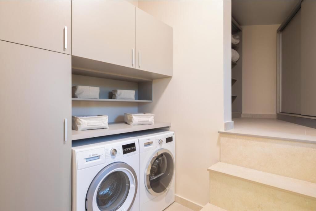 A laundry room with a front-loading washer and dryer, shelving with folded towels, and a staircase leading to a closet.