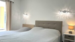 Una cama doble con cabecera capitonada, dos lámparas de pared, una mesita de noche con lámpara, y una ventana con cortinas.