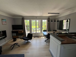 Ein modernes Wohnzimmer mit Flachbildfernseher, einem Essbereich mit Tisch und Stühlen, einer Küche mit Spüle und Arbeitsplatte sowie großen Fenstern mit Vorhängen.