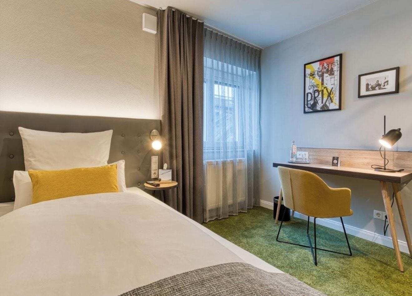 Une chambre d’hôtel comprenant un lit, un bureau, une chaise, une lampe, une fenêtre avec des rideaux et un mur orné d’un tableau.
