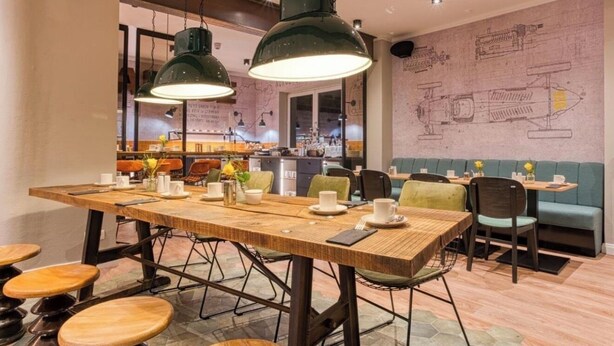Un restaurante con una larga mesa de madera, sillas, lámparas colgantes y una pared con planos técnicos.