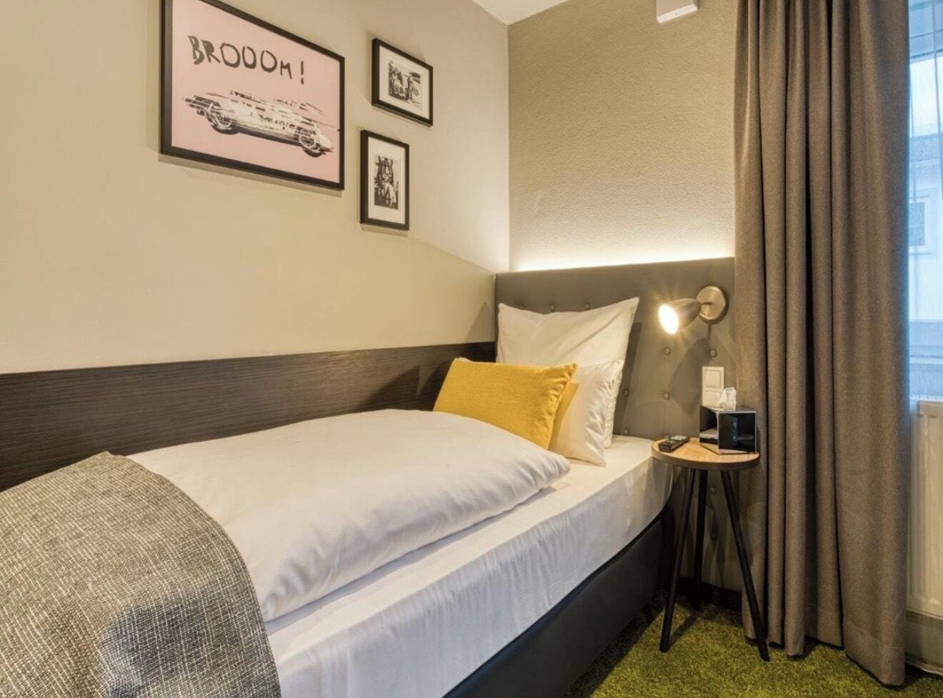 A hotel room with a bed, a bedside table with a lamp, a framed picture on the wall, and a window with curtains.