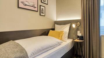 Habitación de hotel con una cama, una mesita de noche con lámpara, un cuadro enmarcado en la pared y una ventana con cortinas.