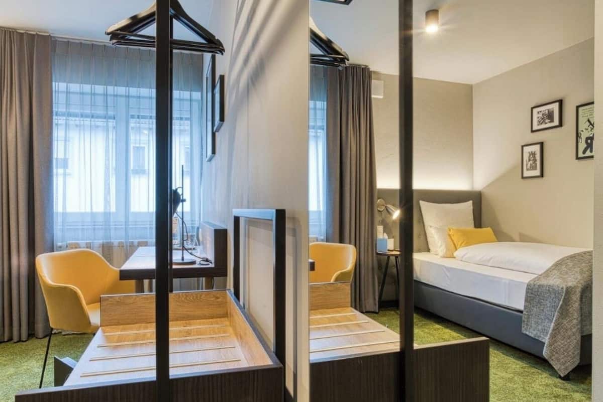 Habitación de hotel con cama, escritorio, dos sillas, ventana con cortinas y pared con cuadros.