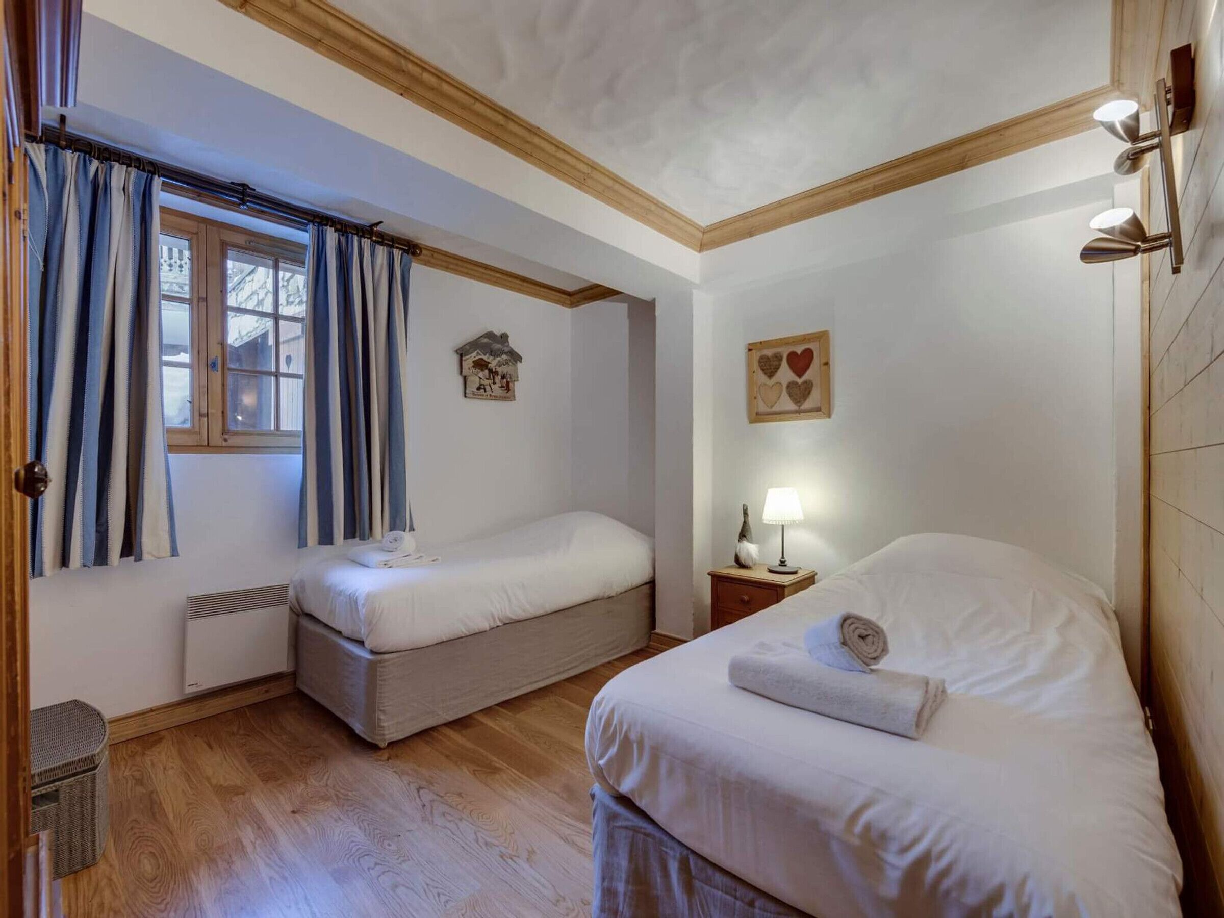 A bedroom with two single beds, a window with curtains, a wall-mounted light fixture, and a small bedside table with a lamp.