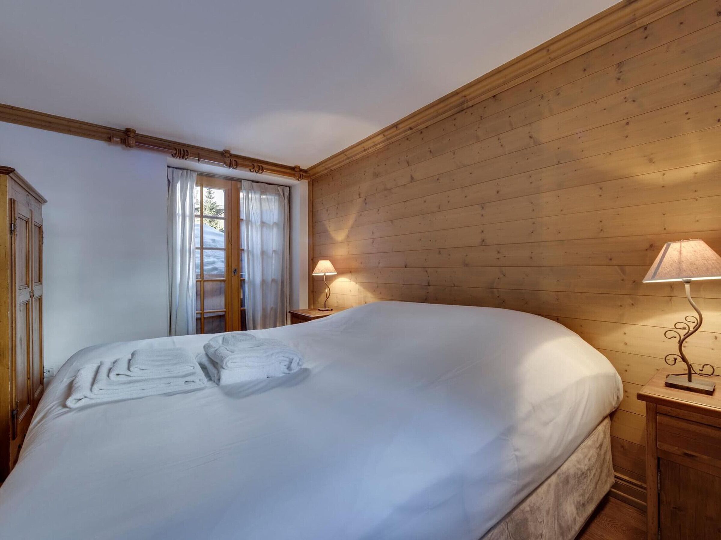 A cozy bedroom with a large bed, wooden walls, a window with curtains, and a bedside lamp.