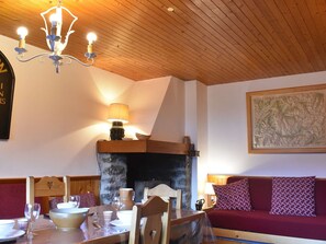 Ein gemütlicher Raum mit Holzbalkendecke, einem Steinkamin, einem Esstisch gedeckt mit Tellern und Gläsern, einer Couch mit gemusterten Kissen und einer Karte an der Wand.