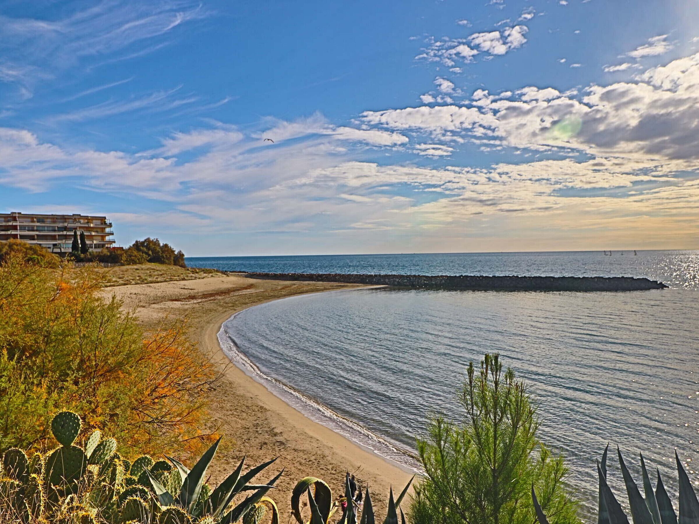 Un paisaje costero con una playa arenosa, un rompeolas rocoso y un edificio con balcones con vistas al mar.