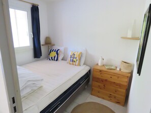 A bedroom with a bed, a wooden nightstand, a window with curtains, and a wall-mounted shelf. - T2 cabin apartment with AC, garden & parking (Sète)